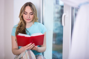 güzel genç kadın kitap ile cam kenarında oturan pijama