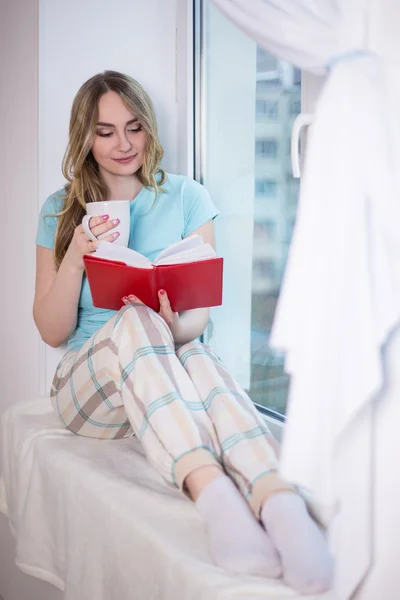 Hermosa mujer joven en pijama sentada junto a la ventana con el libro — Foto de Stock