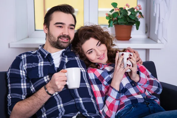 Lyckliga unga paret sitter på soffan med koppar te eller kaffe — Stockfoto