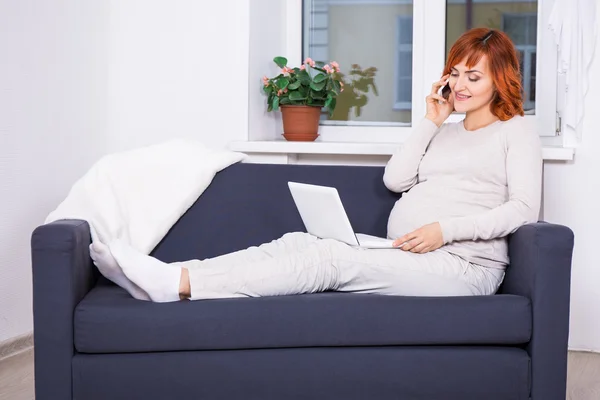 Jovem mulher grávida usando laptop e falando por telefone em vida — Fotografia de Stock