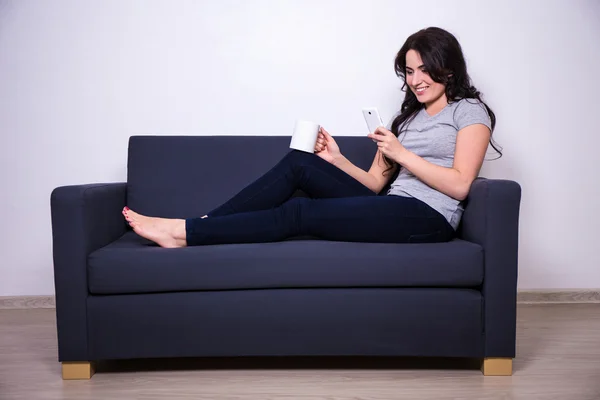 Happy woman sitting on sofa with mobile phone and mug of tea — Stock fotografie
