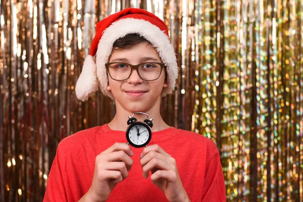 Portrait of a boy with glasses and in a Santa hat with a clock indicating the time on a golden background. Christmas decoration. Twelve Oclock midnight. Clock displays just before New Year. — Stock Photo, Image