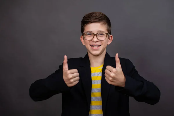 Menino engraçado feliz com óculos mostrar um polegar para cima e boa sorte. em um fundo preto, cinza escuro. — Fotografia de Stock