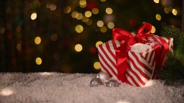 Kırmızı kurdeleli kırmızı ve beyaz çizgili Noel hediyesi karlı yüzeyde gümüş disko topu süslemelerinin yanında duruyor. Noel ağacının arka plan yumuşak bokeh ışıkları, şenlikli dokunuşlar eklenir.