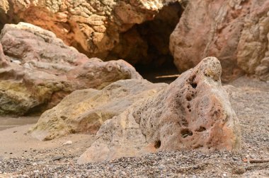 Büyük yıpranmış kıyı kayaları kumlu kıyı şeridinde dinleniyor, sert dokular ve sıcak doğal tonlar sergiliyor. Güneş ışığı taşları plaj boyunca dağılmış halde, erozyonun şekillendirdiği detaylı yüzey şekilleri sergilerler.