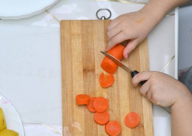 Çocuk güvenli görevlerle bağımsızlığını kazanırken, mutfakta Montessori tarzı öğrenme. Pratik yaşam becerilerinin geliştiğini gösteren eğitici bir an. Çocuk mutfak bıçağıyla haşlanmış havuç kesiyor.