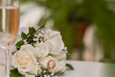 Beyaz güller ve küçük narin detaylarla güzel bir gelin buketi yakın plan. Masaya bir buket çiçek, hafif ışıklandırma, alyanslar, bir bardak şampanya. romantik düğün geçmişi.