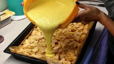 Elma dilimli kek hazırlama, meyve katmanına dökülen pasta hamuru, mutfakta pişirme süreci, ev yapımı tatlı konsepti, taze malzemeler, fırın pişirme aşamasından önce oluşan pasta dokusu.