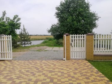 Beyaz çitin ötesinde çakıl yolu görünüyor. Kır manzaralı, kır evi kapısı olan. Kapının dışında nehir var. Ağaçlar ve kırsal çevre kırsal huzuru vurguluyor. Kapıyı aç