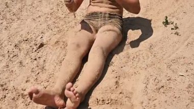 Çocuk güneş ışığında sıcak kumda oturuyor, kumu cilt ve kıyafetlerle örtüyor. Erkek çocuk kumlu arka planda, yaz tatilinde, doğal dokuda, yaşam tarzı konseptinde, doğada sakin boş vakitlerde eğleniyor.