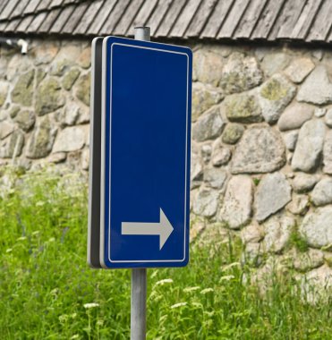 Açık havada taş duvarın yanına yerleştirilmiş beyaz oklu mavi yol işareti. Binanın yanında sağ oku gösteren basit navigasyon işareti, açık ve kolay anlaşılır.