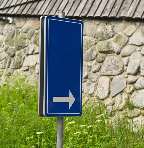 Açık havada taş duvarın yanına yerleştirilmiş beyaz oklu mavi yol işareti. Binanın yanında sağ oku gösteren basit navigasyon işareti, açık ve kolay anlaşılır.