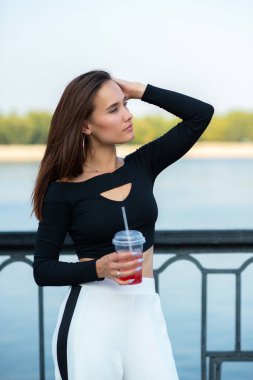 Çekici esmer kız gülümsüyor ve yaz aylarında soğuk bir şeyler içiyor. Genç bir kadının portresi. Doğada nehir, sahil ve mavi temiz gökyüzü ile poz veren kadın model..