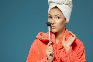 Genç ve çekici bir kadın mavi arka planda elmacık kemiklerine allık sürüyor. Çekici kız sabah güzellik ve makyaj yapıyor.