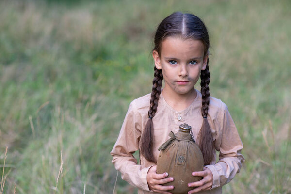 Красивая маленькая девочка с длинными косичками и красивым грязным лицом в летнем лесу и лугу с винтажной фляжкой. Сирота, дитя войны, нищета, голод, голод, бедный бедный ребенок. 