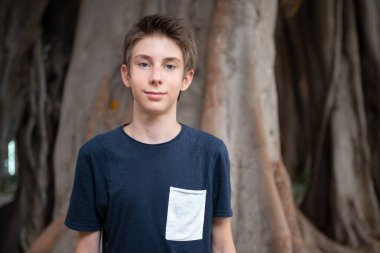 Küçük çocuk incir ağaçlarıyla yaz parkında poz veriyor. Şirin, gözlüklü, gülümseyen, 13 yaşında, köklerinden kameraya bakan genç bir çocuk. Çocuğun açık hava portresi..