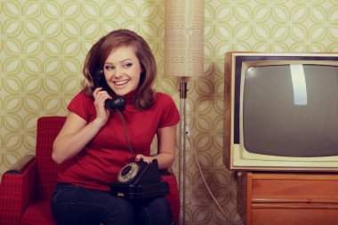Young charming woman sitting in chair and talking on old phone in room with vintage wallpaper and retro TV set, retro stylization 60-70s 20th century. Girl in room with old fashioned interior clipart
