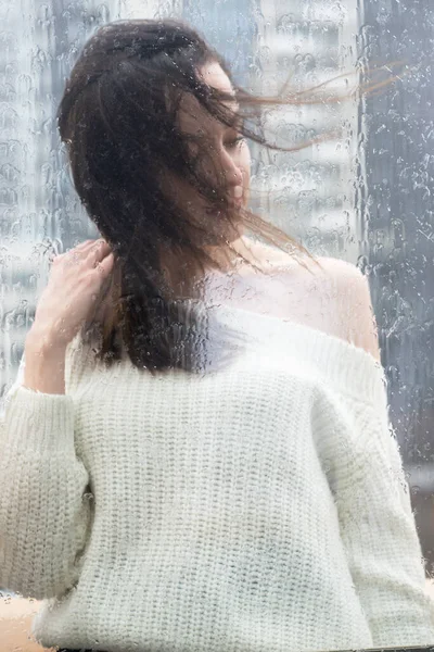 年轻美丽的女人 一头乌黑的长发 穿着一件白色的毛衣 窗外的窗玻璃后面是雨滴 在家里的肖像 自我隔离 待在家里 隔离观念 — 图库照片