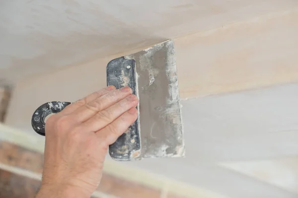 Worker putsty plasterboard ceiling in new building. Repairman works with plasterboard, plastering dry-stone wall, home improvement. A man makes repairs at home. Putty knife in male hand