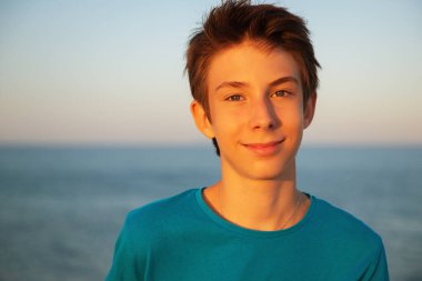 Handsome young happy boy enjoing life at beach at sunset. Beautiful tan calm smiling teen boy wearing blue t-shirt posing alone at Mediterranean sea coast. Travel, summer vacation, tourism, teenage lifestyle.