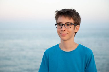 Kumsalda, güneşin doğuşunda ya da batışında yakışıklı, mutlu, gözlüklü çocuk. Akdeniz kıyısında yalnız poz veren güzel, sakin, gülümseyen genç bir çocuk. Seyahat, yaz tatili, turizm, gençlik yaşam tarzı.