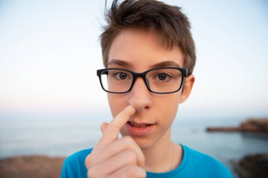 Komik bir çocuk burnunu karıştırıyor, kameraya bakan bir şey düşünüyor, gün batımında sahile bakıyor, geniş açıdan. Bronz tenli, gözlüklü, mavi tişörtlü, Akdeniz kıyısında yalnız poz veren genç bir çocuk.. 