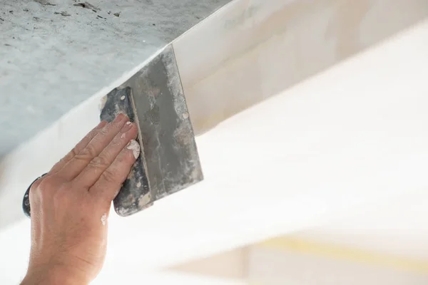 Worker putsty plasterboard ceiling in new appartment. Repairman works with plasterboard, plastering dry-stone wall, home improvement. A man makes repairs at house. Putty knife in male hand. Hand work