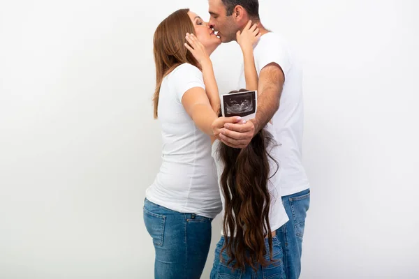 怀孕了快乐的家庭期待宝宝和显示超声扫描的孩子在肚子里 男人抱着一个大肚子的孕妇 亲吻她 妻子与女儿穿着休闲装 蓝色牛仔裤和白色T恤 的丈夫的室内肖像 — 图库照片