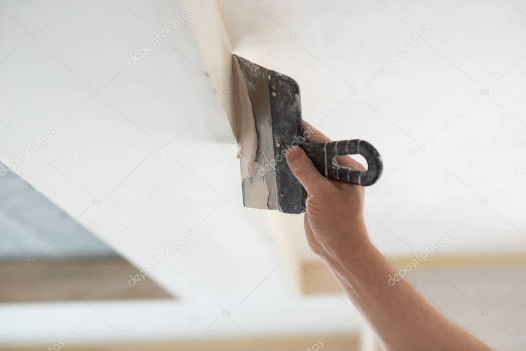 Worker putsty plasterboard ceiling in new building. Repairman works with plasterboard, plastering dry-stone wall, home improvement. A man makes repairs at home. Putty knife in male hand
