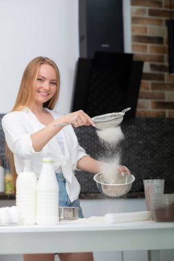 Genç bir kadın mutfakta duruyor ve un, süt, kakao, şeker ve meyvelerden oluşan fırın ürünleriyle yemek pişiriyor. Mutlu bir kızın komik portresi. Şef un süzüyor.