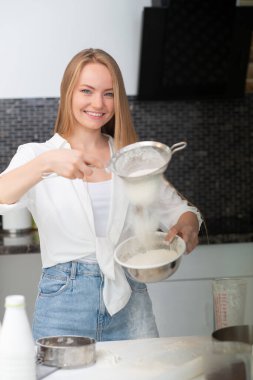 Genç bir kadın mutfakta duruyor ve un, süt, kakao, şeker ve meyvelerden oluşan fırın ürünleriyle yemek pişiriyor. Mutlu bir kızın komik portresi. Şef un süzüyor.