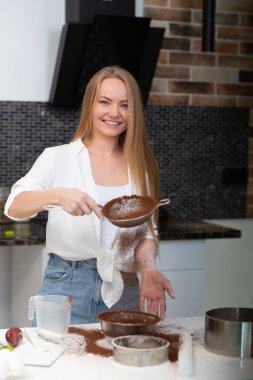 Genç bir kadın mutfakta duruyor ve un, süt, kakao, şeker ve yumurtalardan oluşan fırın ürünleriyle yemek pişiriyor. Mutlu kız şefin komik portresi. 