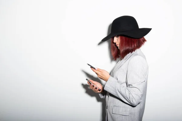 スマートフォンを使用して若いスタイリッシュな女性 白の上に屋内の肖像画 ファッション現代女の子身に着けていますグレージャケットと黒帽子テキストメッセージ上の彼女の携帯電話 — ストック写真
