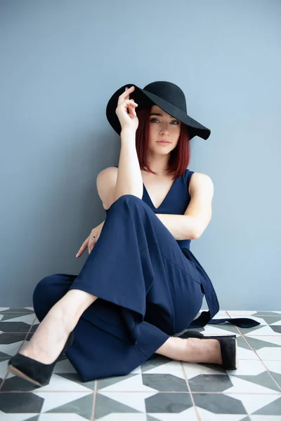 深い青の服と黒の帽子を身に着けているファッション女性は床に座って屋内をポーズ — ストック写真