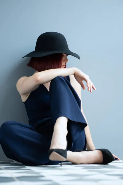 深い青の服と黒の帽子を身に着けているファッション女性は床に座って屋内をポーズ — ストック写真