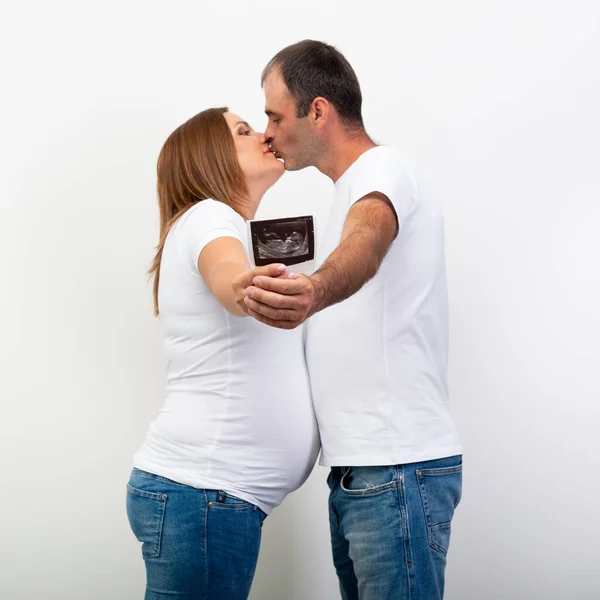 怀孕了快乐的家庭期待宝宝和显示超声扫描的孩子在肚子里 男人抱着一个大肚子的孕妇 亲吻她 — 图库照片