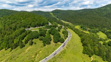 Romanya 'daki Oituz Geçidi' ni geçen, yeşil ormanlık tepeler ve parçalı bulutlu bir yaz gökyüzü altında açık çayırlarla çevrili bir dağ yolunun hava manzarası..