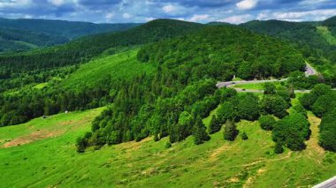 Romanya 'daki Oituz Geçidi' ni geçen, yeşil ormanlık tepeler ve parçalı bulutlu bir gökyüzü altında açık çayırlarla çevrili bir hava aracı videosu..