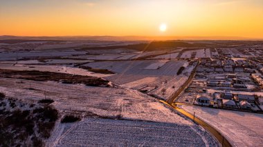 Gündoğumunda, Romanya 'nın Miroslava kentindeki karla kaplı tarım arazileri ve yerleşim bölgelerinin hava manzarası, kayan tarlalar, kırsal yollar ve sakin bir manzaranın üzerindeki yumuşak altın kış ışığı.