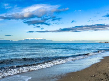 Yunanistan 'ın kuzeyindeki Chalkidiki' de kumlu bir plajdan Ege Denizi boyunca karla kaplı Olimpos Dağı 'nın sabah manzarası. Dalgalar kıyıya ulaşır mavi gökyüzünün altında ve dağınık bulutlar uzak dağ manzaralı sakin bir kıyı manzarası yaratır.
