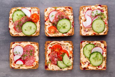 Krem peynirli, sebzeli ve salamlı sandviç. Salatalıklı sandviçler, turp, domates, gri arka planda salam, üst manzara. Düz yatıyordu.