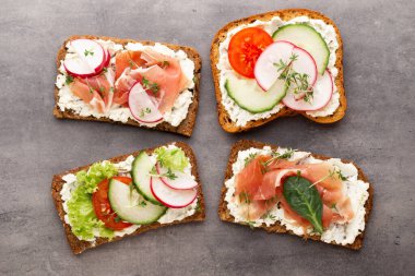 Krem peynirli, sebzeli ve salamlı mini sandviçler. Salatalıklı sandviçler, turp, domates, gri arka planda salam, üst manzara. Düz yatıyordu.