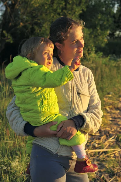 Jeune jolie jeune mère tenant une petite fille dans ses bras au coucher du soleil. Promenade en automne soirée chaude en plein air au coucher du soleil . — Photo