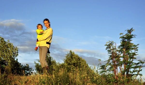 Joven atractiva joven madre sosteniendo a una niña en sus brazos al atardecer. Camine en la cálida noche de otoño al aire libre al atardecer . — Foto de Stock