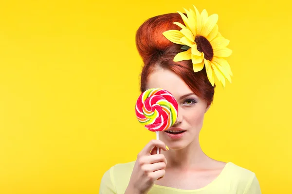 Bella ragazza con una grande caramella, isolata su uno sfondo giallo con fiore giallo nei capelli, emozioni, cosmetici — Foto Stock