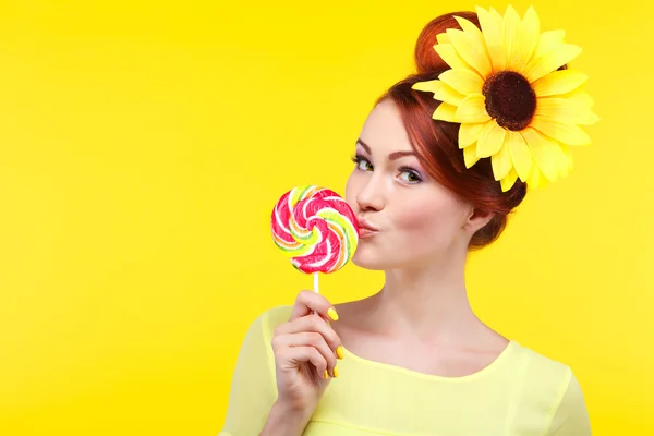 Hermosa chica con un gran caramelo, aislado sobre un fondo amarillo con flor amarilla en pelos, emociones, cosméticos —  Fotos de Stock