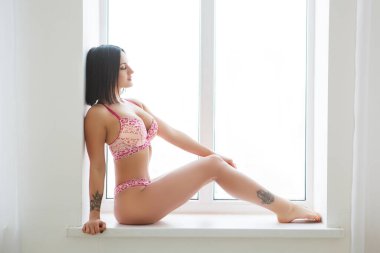 Pencereye poz veren seksi esmer güzel. Seksi esmer kadın iç çamaşırları. Seksi iç çamaşırlı güzel romantik bir kadının portresi. Arka planda dinleniyor..