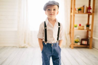 Beyaz gömlekli, şapkalı ve askılı parlak bir odada tatlı bir çocuk. Giysilerdeki Retro biçimi