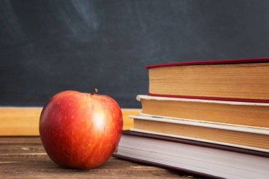 Tahta ya da karatahtanın arka planına karşı ahşap bir masanın üzerinde kitaplar ve elmalar.