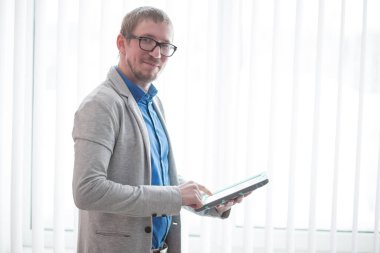 Ofisteki büyük pencereli genç adamın portresi. Tablet bilgisayar kullanan ve kameraya bakan adam.